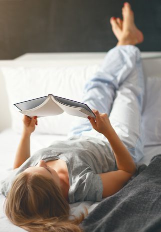 Happy young woman reads book and drinks coffee in bed